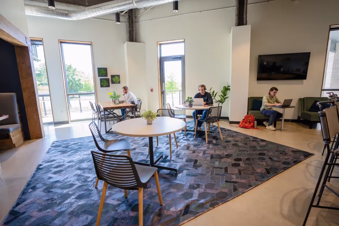 A modern, bright coworking space with large windows, tables, chairs, a patterned rug, and three people working on laptops. Green plants and wall art decorate the space; a red backpack is on the floor.