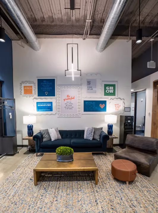 Modern lounge area with a dark blue sofa, brown chair, wooden coffee table with a plant, two blue lamps, and motivational posters beneath exposed ceiling pipes—perfect for coworking space or event spaces in Columbus, Ohio.
