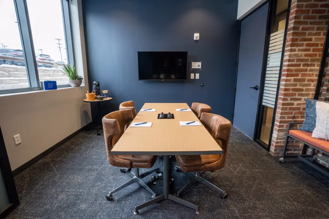A small modern conference room with a rectangular table, four brown chairs, notepads with pens, a wall-mounted TV, and large windows letting in natural light. Perfect for private offices or meeting rooms, with a coffee station on a side counter.