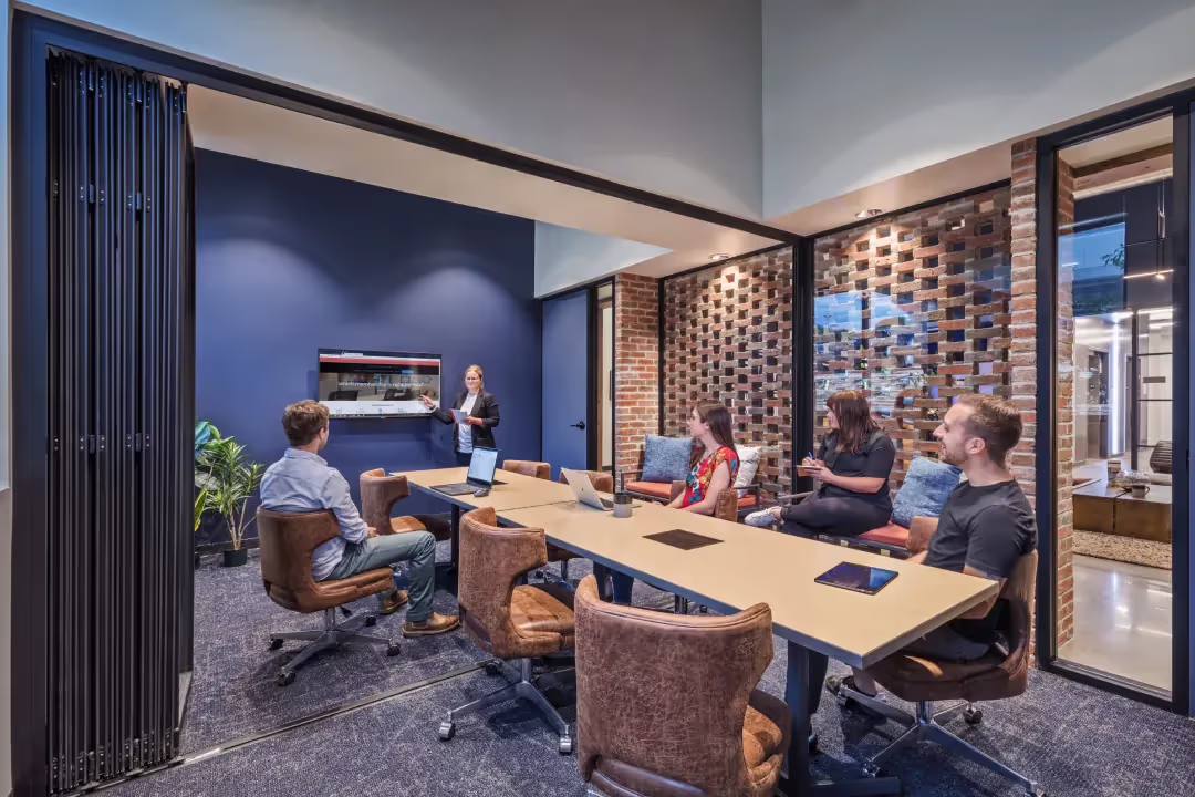 Five people are in a bright, collaborative conference room with brick and glass walls—perfect for teams seeking office space for rent. Four are at a long table with laptops, while one presents by the screen, highlighting a flexible workspace atmosphere.
