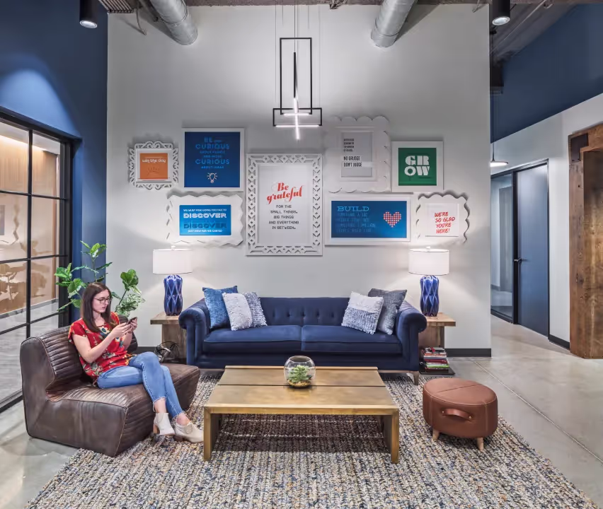 A woman sits on a leather chair using her phone in a modern lounge with a blue sofa, two table lamps, a wooden coffee table, and colorful framed quotes—an inviting office space for rent with industrial-style decor.