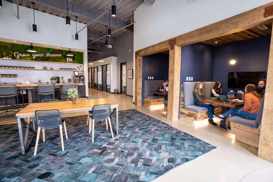 Modern coworking space with open seating, a wooden table, blue-patterned rug, kitchen area, and booth seating where people are working and talking. A sign on the wall reads “Grow your dreams.” Meeting rooms are also available.