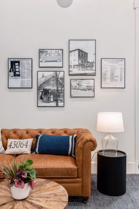 A cozy living room with a tan tufted sofa, a blue pillow, a round wooden coffee table with a potted plant—perfect for relaxing or brainstorming in your coworking space—and six framed black-and-white photos and articles on the white wall.