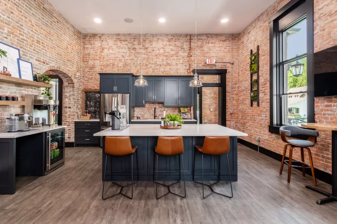 Modern kitchen with exposed brick walls, large windows, dark blue cabinets, a white island with three brown chairs, pendant lights, wood flooring, and plants on the counter—perfect for a coworking space or casual meetings.