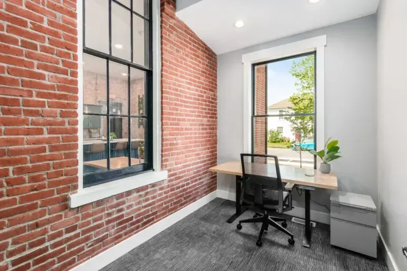 A small coworking space with exposed brick walls, a modern desk and chair, a potted plant, a window overlooking a tree and building, plus carpeted flooring—ideal for focused work or as a virtual business address.