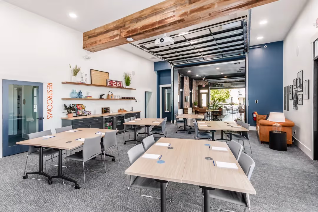 Modern meeting room in a coworking space with wooden tables, gray chairs, notepads, and pens. Enjoy a lounge, kitchenette, digital clock, restrooms sign, and large windows. Ideal for those seeking meeting rooms or event spaces in Columbus, Ohio.