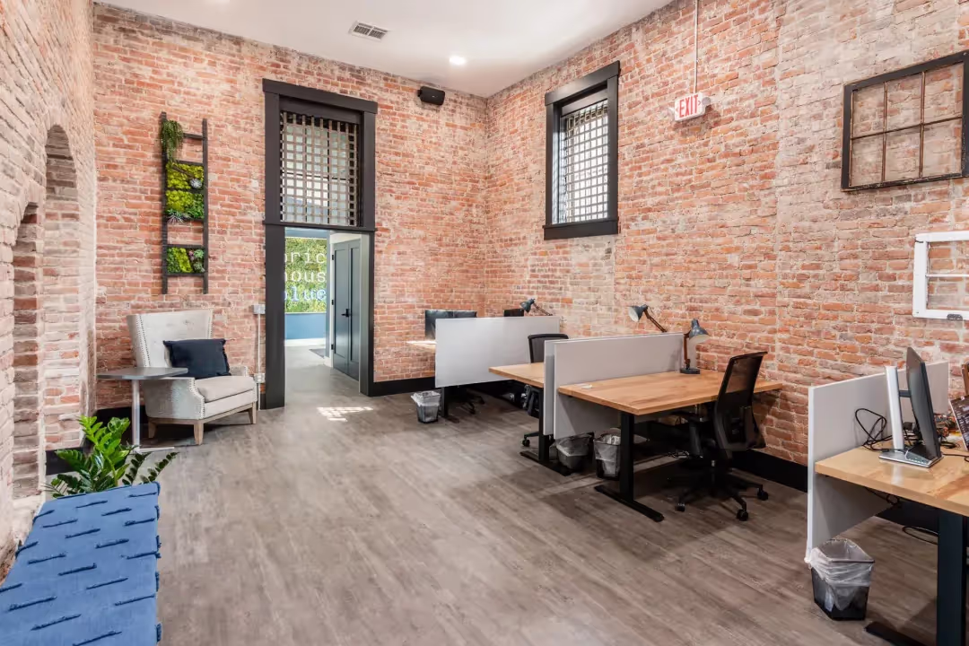 A modern coworking space with exposed brick walls, desks with computers, gray chairs, and a wooden floor. There is a blue bench, a lounge chair, some plants, and a large window letting in natural light.