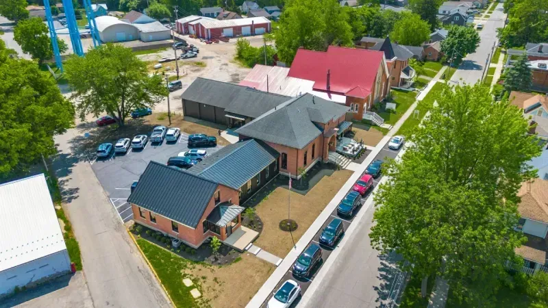 Aerial view of a small town street with brick buildings, parked cars, green trees, and residential houses—some offering private offices and office space for rent—under a partly cloudy sky.