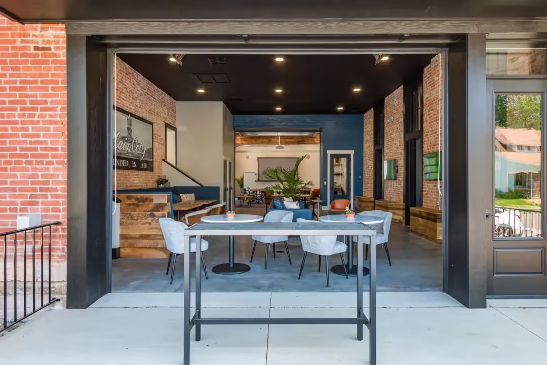 Modern café with an open garage-style door, exposed brick walls, wood and blue accents, and various tables and chairs—ideal as a relaxed coworking space in a welcoming, indoor-outdoor setting.