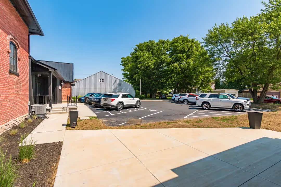A paved patio area overlooks a small parking lot with several parked cars. Trees provide shade in the background, while a brick building on the left features modern coworking space and meeting rooms under a clear blue sky.