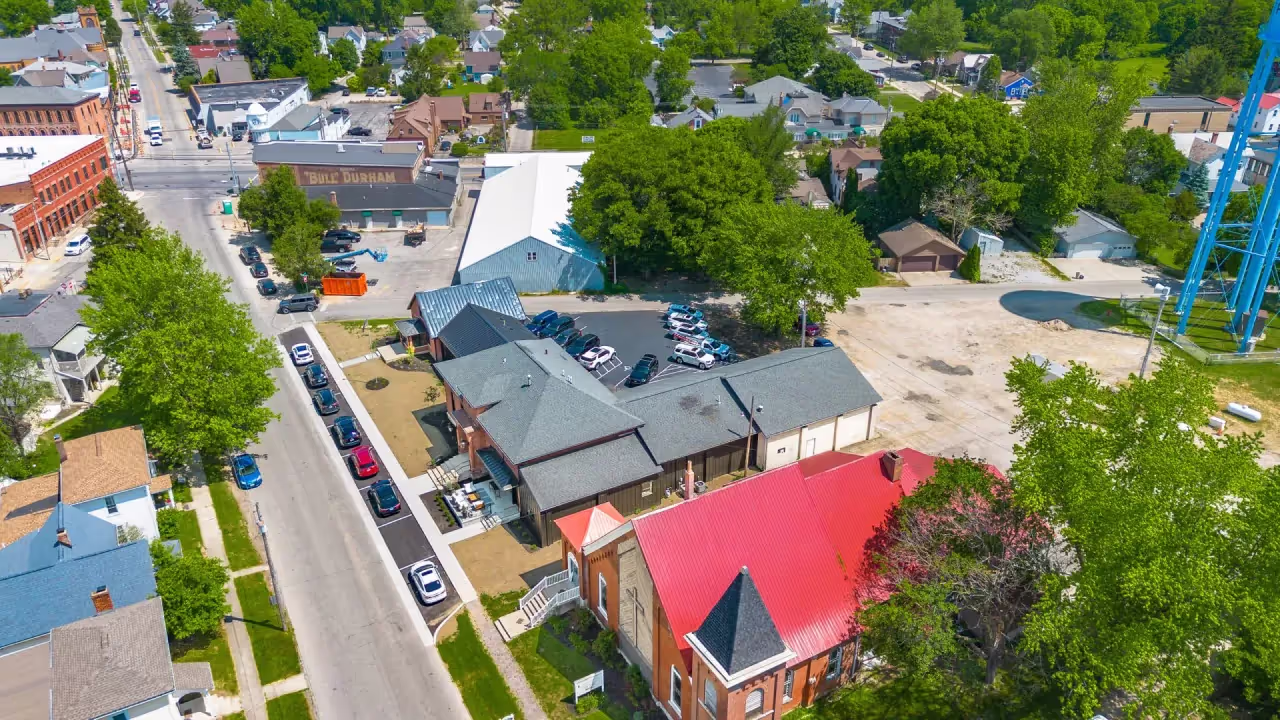 Aerial view of a small town with houses, parked cars along a street, a red-roofed building offering meeting rooms, trees, and a blue water tower in the background.