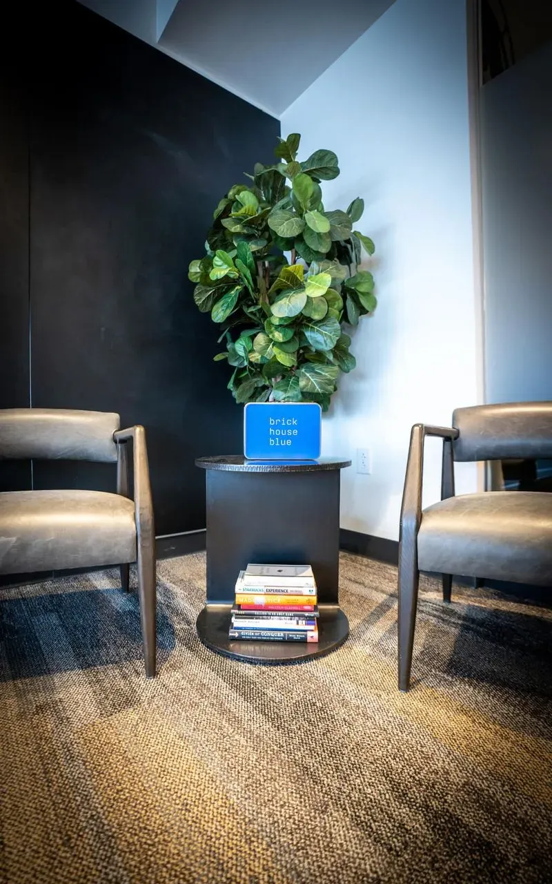 Two gray chairs sit on either side of a small black table with a potted plant and a blue sign on top, creating a cozy nook in this coworking space. Several stacked books rest on a lower shelf. The room has carpeted flooring and neutral walls.