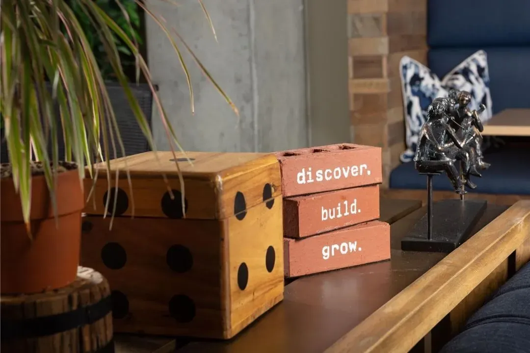 A wooden table in a coworking space with a potted plant, large wooden dice, three bricks labeled discover., build., grow., and a small sculpture of people sitting together. Ideal for meeting rooms or event spaces Columbus Ohio.