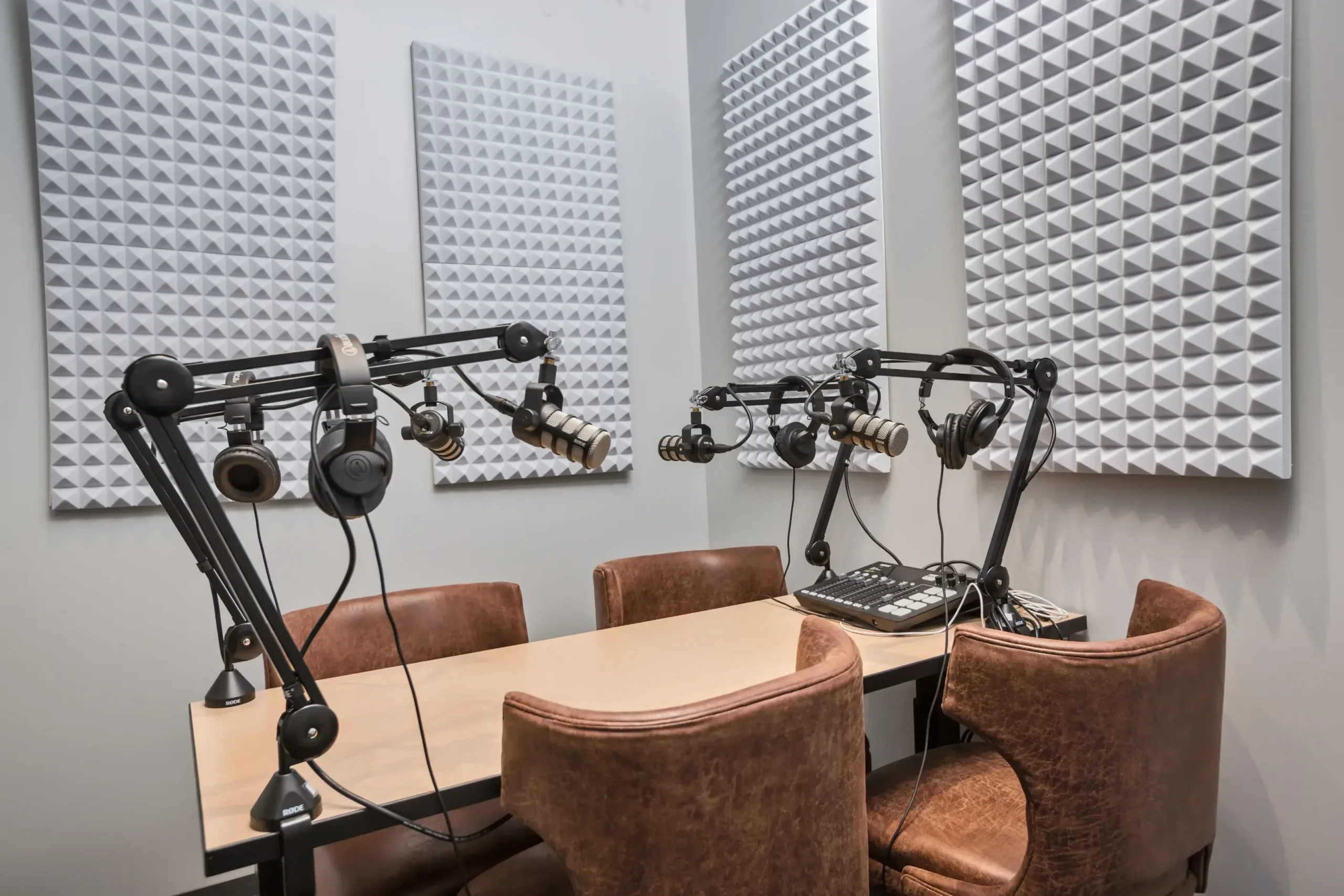 A modern podcast studio in a coworking space with four brown chairs around a table, microphones on adjustable arms, headphones, and soundproof foam panels on the walls for acoustic treatment.