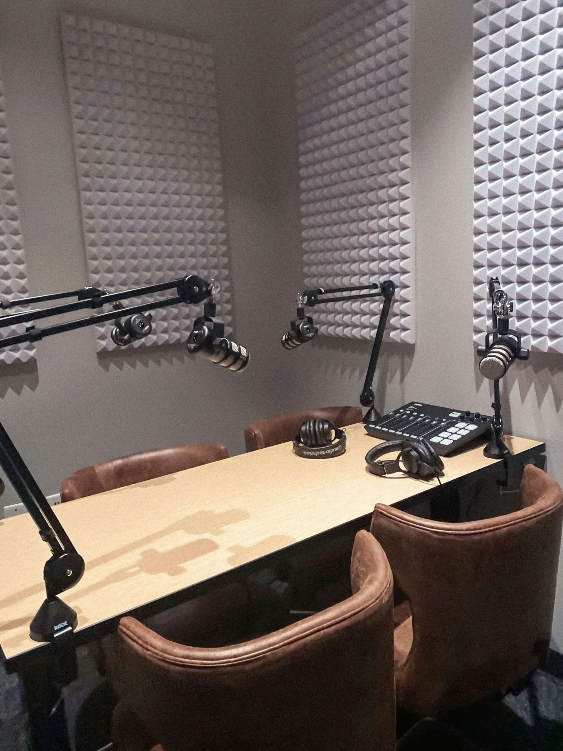 A podcast recording studio with four microphones on adjustable arms, soundproof foam panels, and a wooden table surrounded by brown chairs—plus access to private offices and meeting rooms for all your creative needs.
