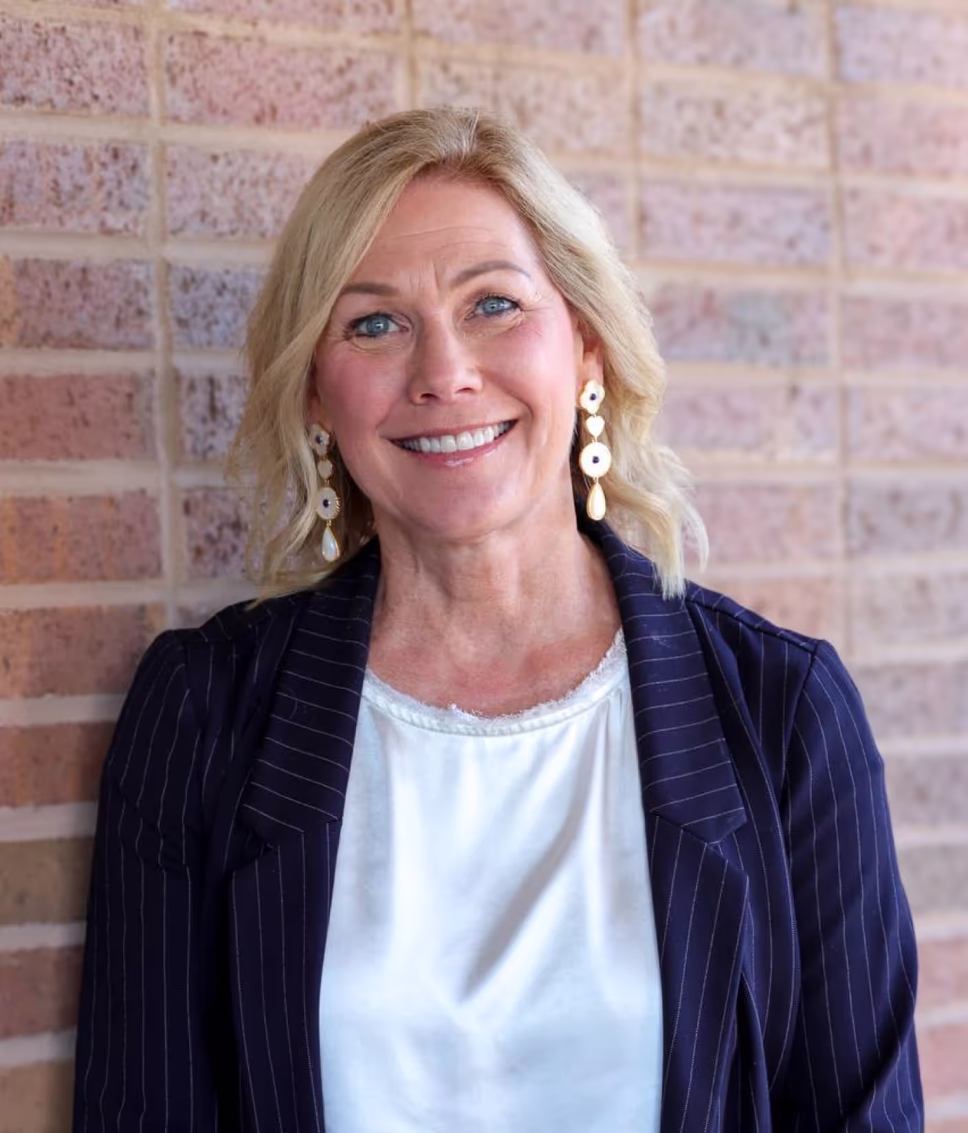 A smiling woman with blonde hair wearing a white blouse, dark pinstripe blazer, and dangling earrings stands in front of a brick wall, embodying the professional vibe of modern office space for rent and private offices.