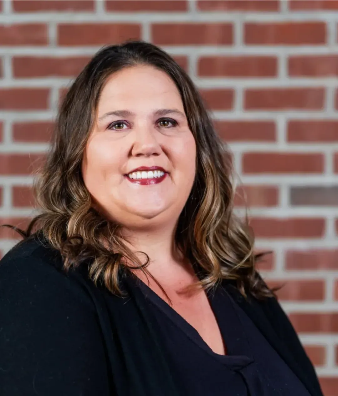 A woman with wavy brown hair, wearing a black top, smiles at the camera while standing in front of a red brick wall—an inviting backdrop often found in stylish coworking spaces or private offices in Columbus, Ohio.