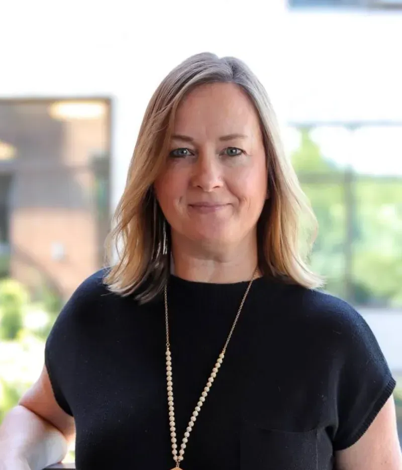 A woman with shoulder-length blonde hair, wearing a black top and a long beaded necklace, stands outdoors in front of greenery and buildings—possibly near a coworking space. She is smiling gently at the camera.