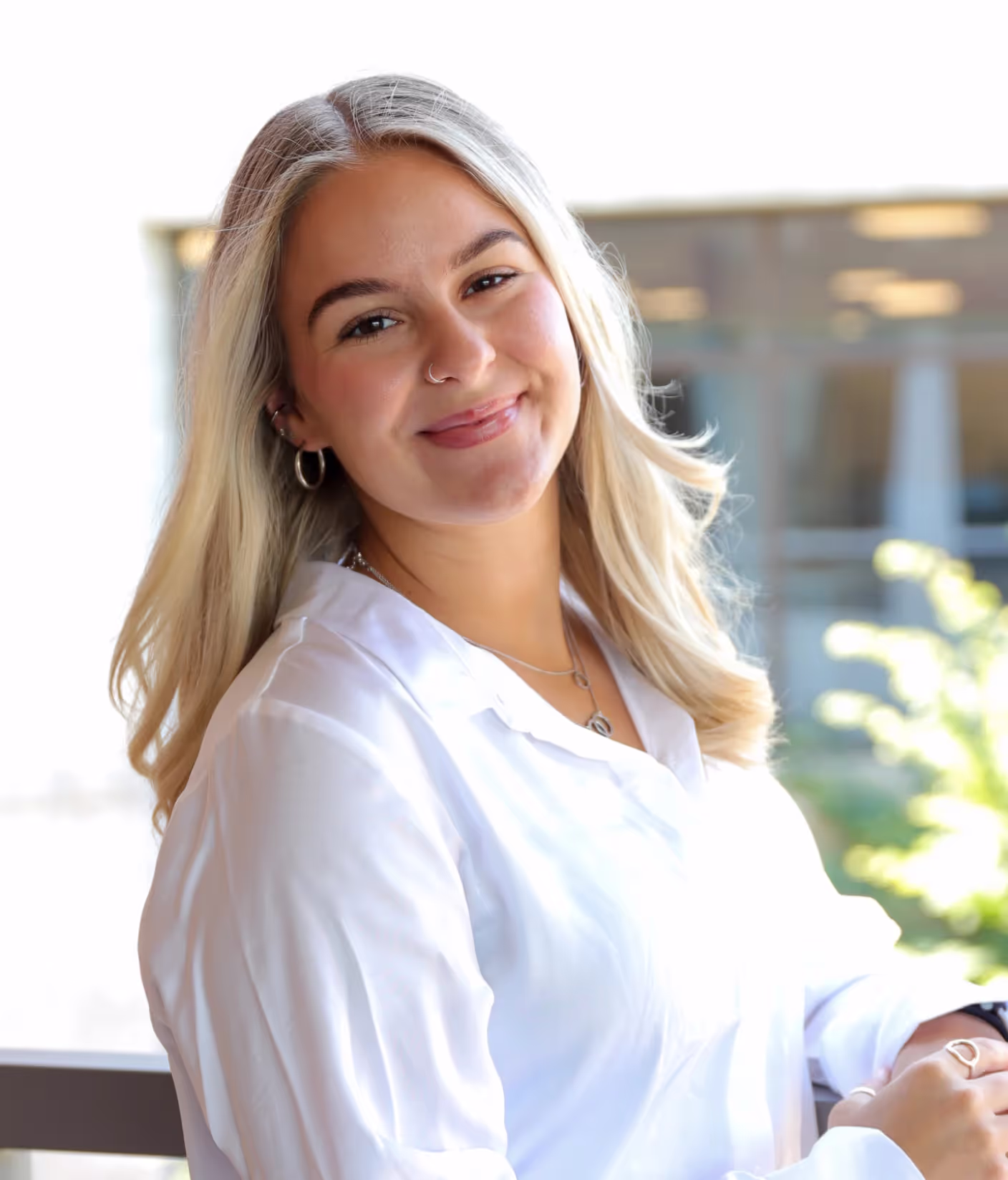 A woman with long, blonde hair and a white blouse smiles at the camera while standing outdoors, with a blurred office space for rent and greenery in the background.