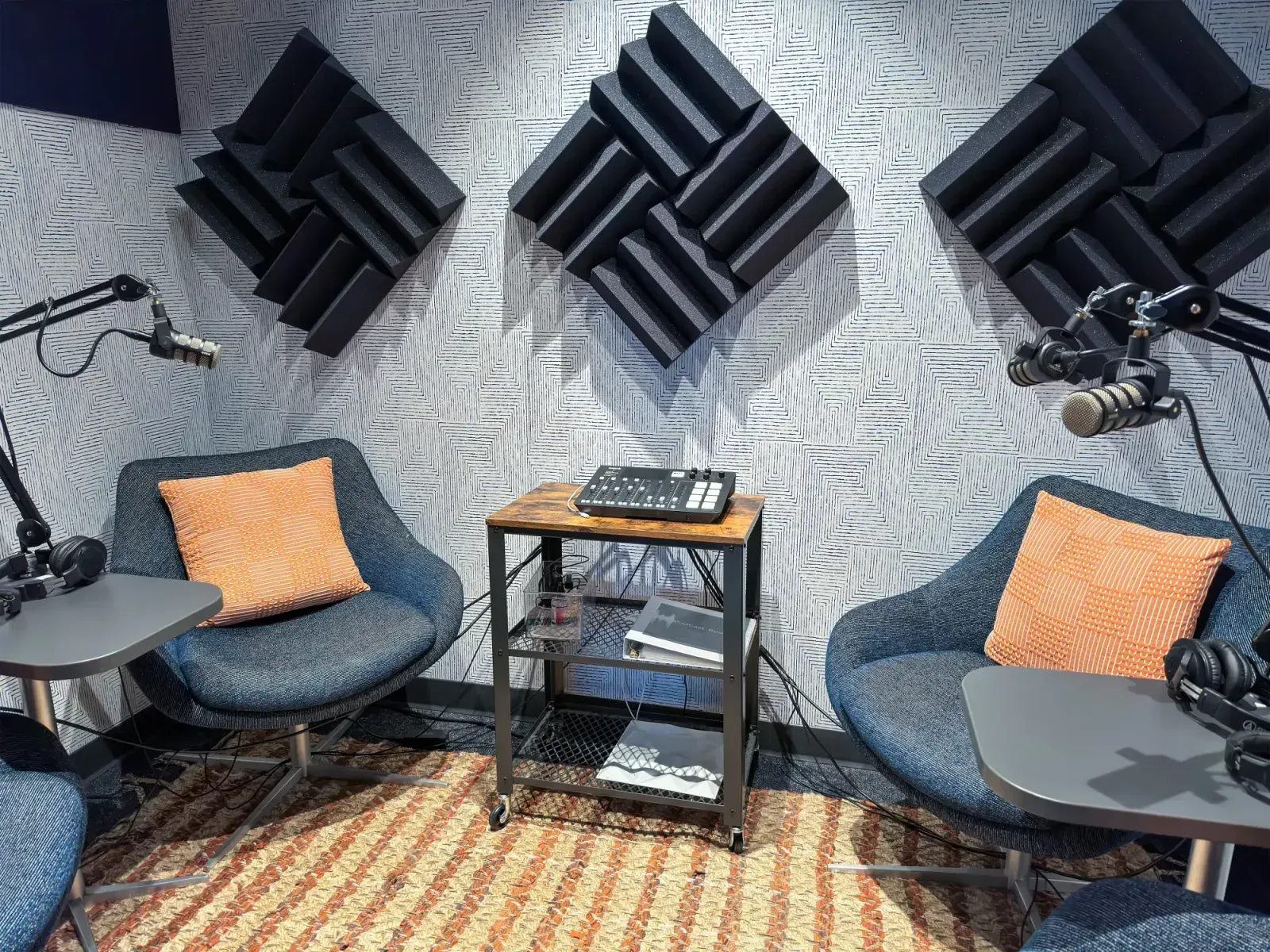 A modern podcast recording studio in a coworking space, featuring two chairs with orange pillows, microphones on adjustable arms, acoustic foam panels on the wall, and audio equipment on a small rolling cart between the chairs.