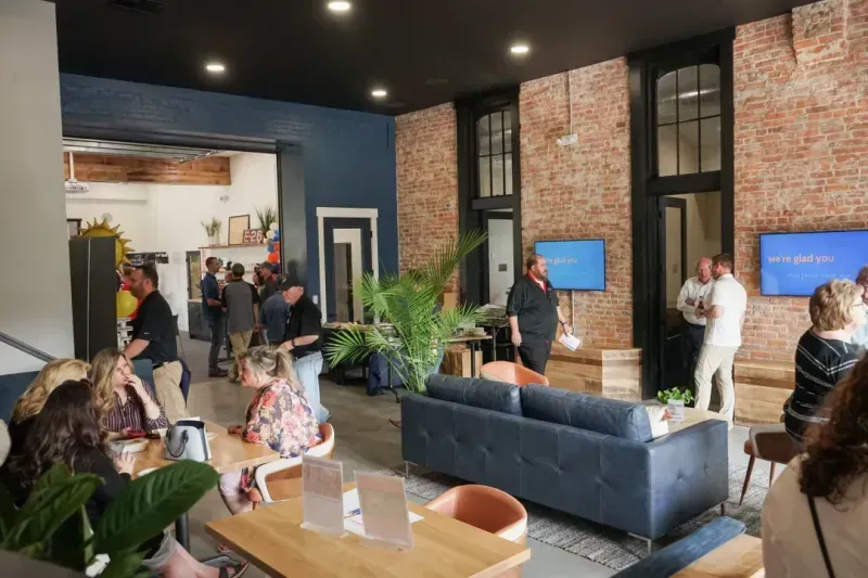 People gather in a modern coworking space with exposed brick walls, blue accents, and large windows. Groups sit at tables or chat near sofas. Two digital screens display the message “we’re glad you’re here.”.