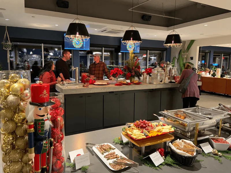 A festive buffet setup with various cheeses, fruits, and hot dishes on a counter, decorated with Christmas ornaments and a nutcracker figure—perfect for gatherings in event spaces Columbus Ohio—while guests serve food and chat in a warmly lit, modern dining area.
