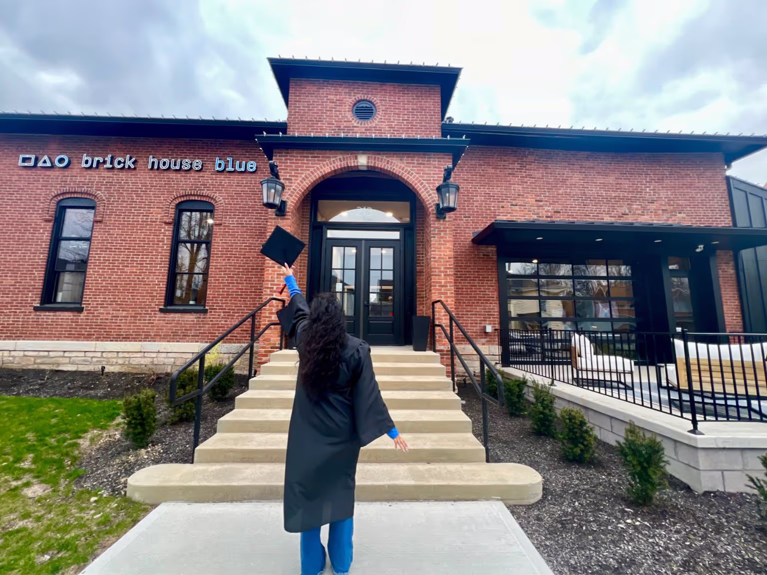 A person in a graduation gown stands on the steps, holding up a cap outside Brick House Blue, known for event spaces Columbus Ohio. The cloudy sky frames the brick building’s large windows and impressive central entrance.
