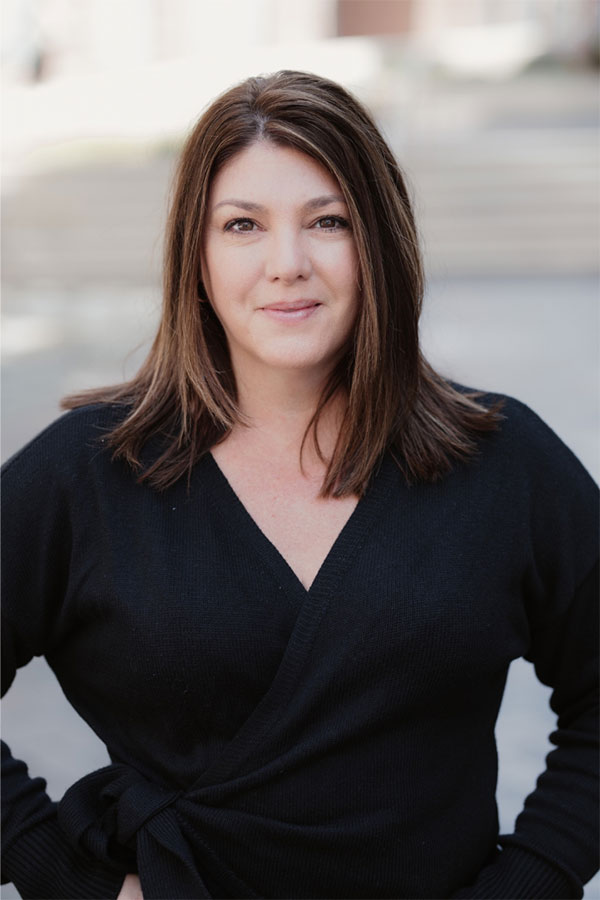 A woman with straight brown hair and light skin wearing a black wrap sweater stands outdoors, smiling softly at the camera. The background is blurred, echoing the focus of a professional seeking meeting rooms or office space for rent.