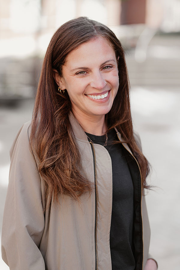 A woman with long brown hair, wearing a light brown jacket over a black top, smiles while standing outdoors in soft natural light—perfectly embodying the professional vibe of private offices or an ideal virtual business address.