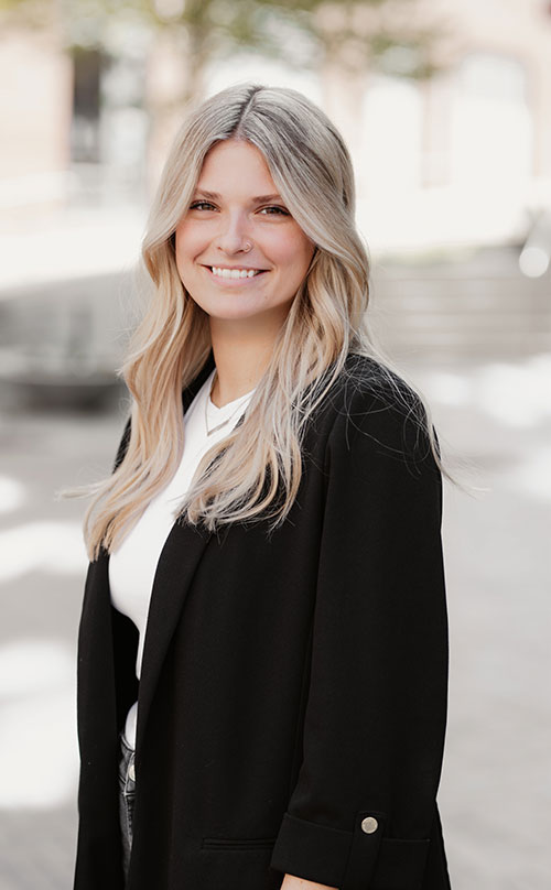 A woman with long, blonde hair wearing a black blazer and white top smiles at the camera while standing outdoors in a sunlit area, perfect for those seeking meeting rooms or office space for rent in a welcoming, professional setting.