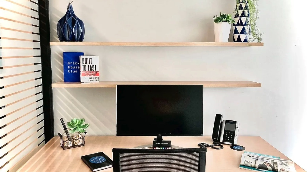 A modern, minimal desk setup ideal for private offices, featuring a monitor, phone, notebooks, plant, and pen holder. Two shelves above hold books, vases, and more plants. A magazine and a coaster sit on the light wood desk.