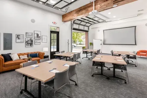 Modern meeting room with several tables and chairs arranged in small groups, a large projector screen, framed photos on the wall, a brown sofa, and large windows letting in natural light—ideal for coworking space or professional meeting rooms.