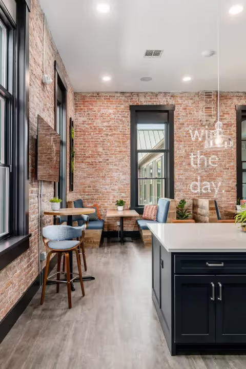 Modern kitchen and lounge area with exposed brick walls, large black-trim windows, a flat-screen TV, a central island, and cozy seating; “win the day” is written on one wall—adjacent private offices and meeting rooms available.