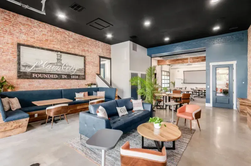 A modern, open-concept lounge features blue sofas, round wooden tables, and stylish chairs on polished concrete floors. Exposed brick walls and plants add warmth, while nearby private offices and a “Palm City” sign complete the inviting space.