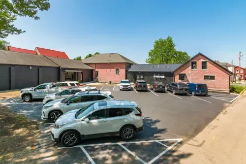 A small parking lot with several parked cars is in front of brick and dark-colored buildings offering office space for rent on a sunny day with clear blue skies. Some trees and houses are visible in the background.