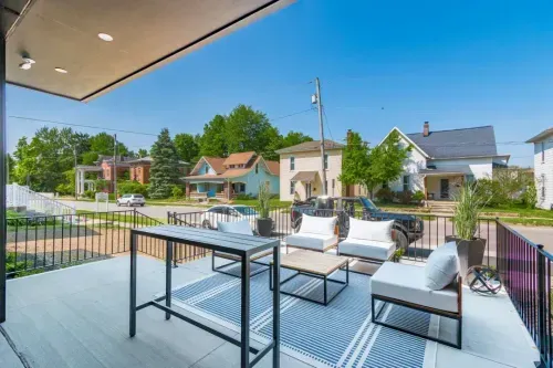 Modern outdoor patio with a table, sofas, and potted plants on a rug—ideal for relaxed meetings or coworking space—overlooking a quiet suburban street with houses and trees under a clear blue sky.