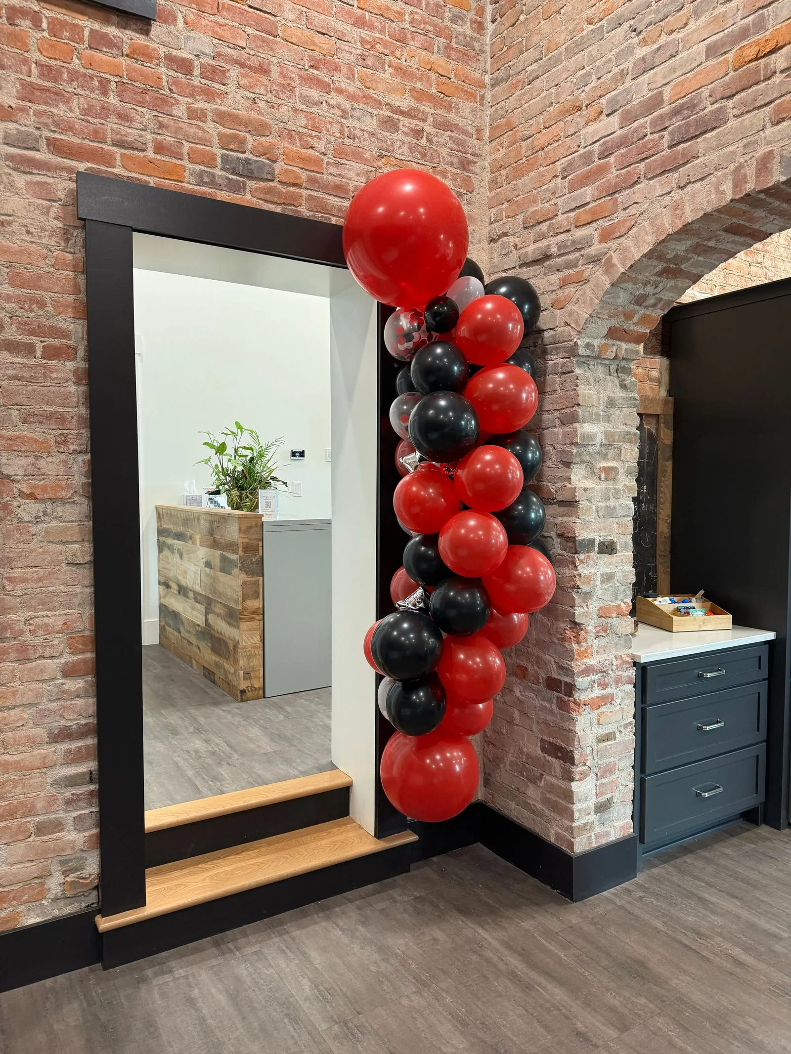 A cluster of red and black balloons is arranged vertically next to a doorway in a room with exposed brick walls and wooden floors, highlighting the inviting charm often found in event spaces Columbus Ohio has to offer. A reception desk with a plant is visible through the doorway.