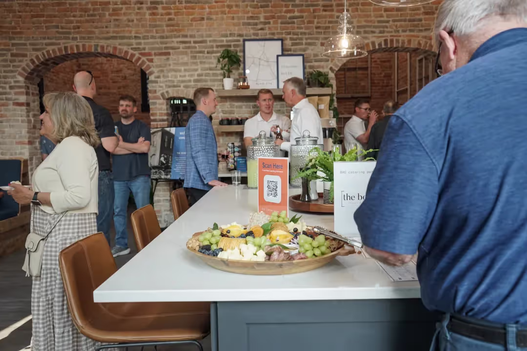 People gather and socialize in a modern space with exposed brick walls. A large table in the foreground holds fruit and cheese, while signs and plants decorate this inviting event space in Columbus, Ohio.