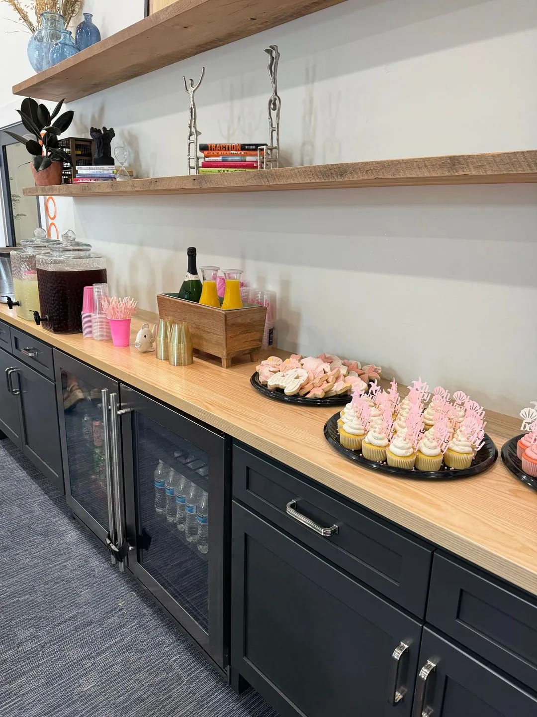 A countertop in a vibrant coworking space features trays of pink cupcakes, cookies, drink dispensers, and champagne bottles; shelves above hold books and decor. Black cabinets below store drinks and supplies—perfect for event spaces in Columbus, Ohio.